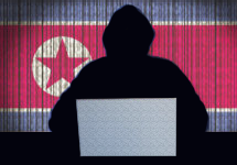 North Korean flag in the background a shadowed figure in a hoodie at a desk with the light of a laptop in front