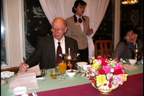 Lewis writing in notepad during a dinner with woman sitting next to him and a waitress standing behind