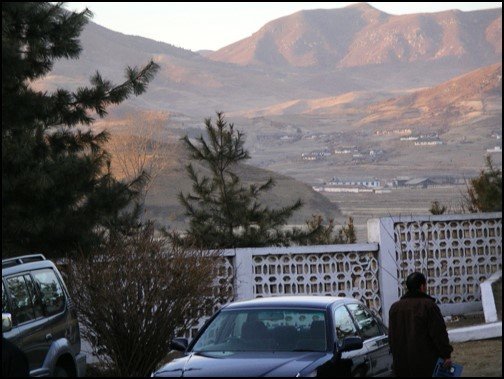 A view of distant mountain range and some low buildings at the foot. Cars and an ornamental concrete fence in the foreground