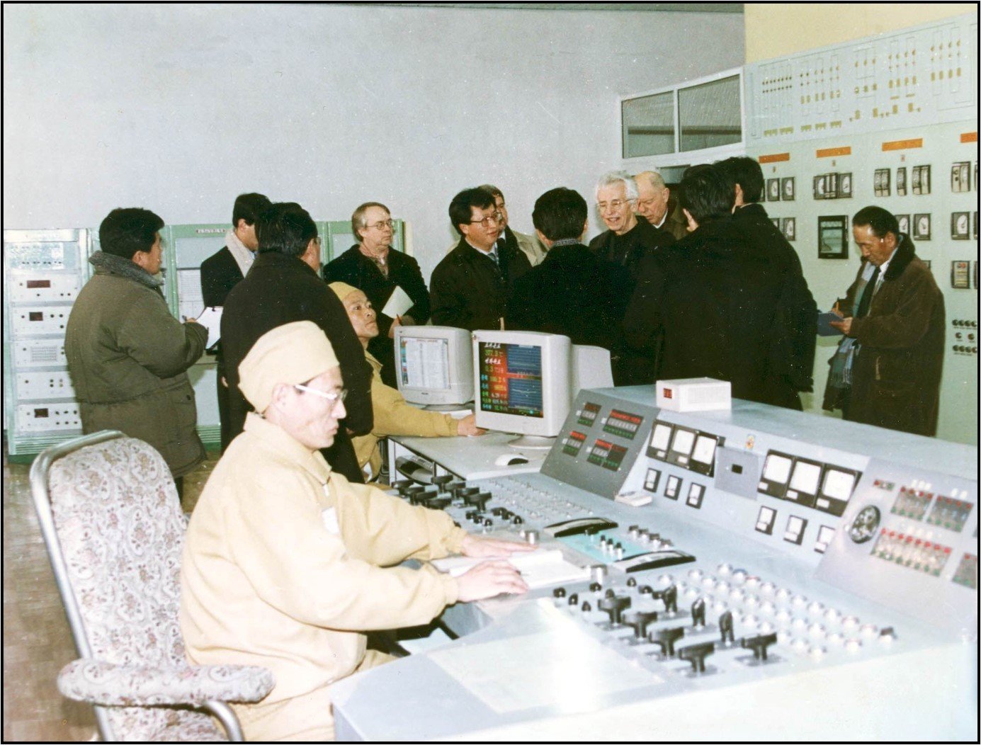 Man in protective clothing sits at the panel of 5 MWe control room while a large group of people talks in the background