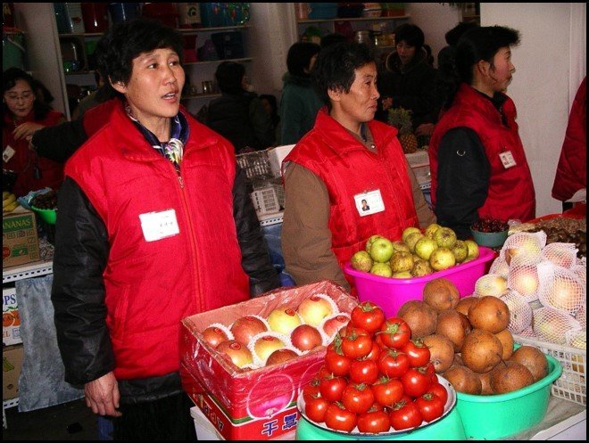Two women wearing red vests and name tags standing behind a display of fruit