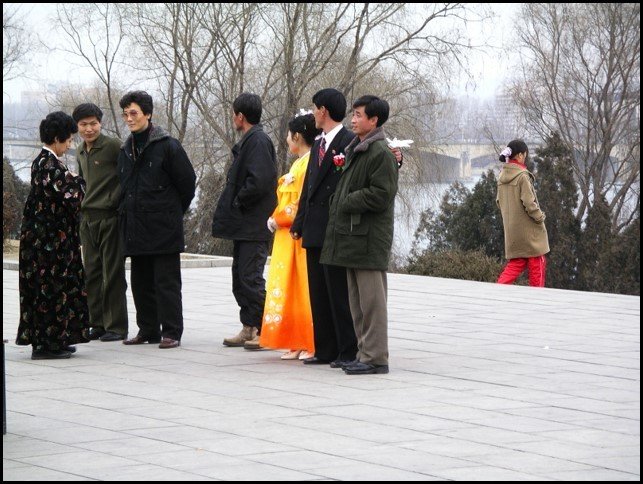 Bride in long orange robe and the groom in traditional suit surrounded by men and being addressed by another woman in a long Korean robe
