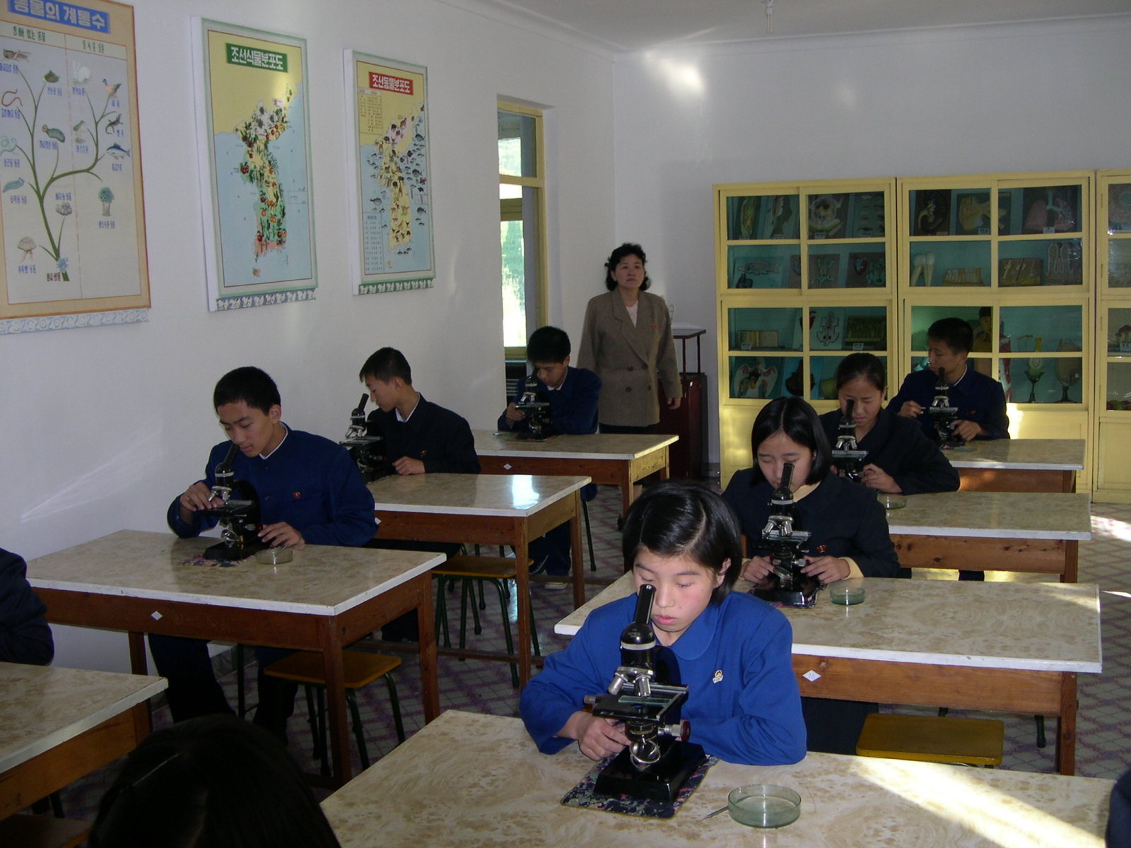 Students sitting at their desks and looking into microscopes with a woman teacher looking on