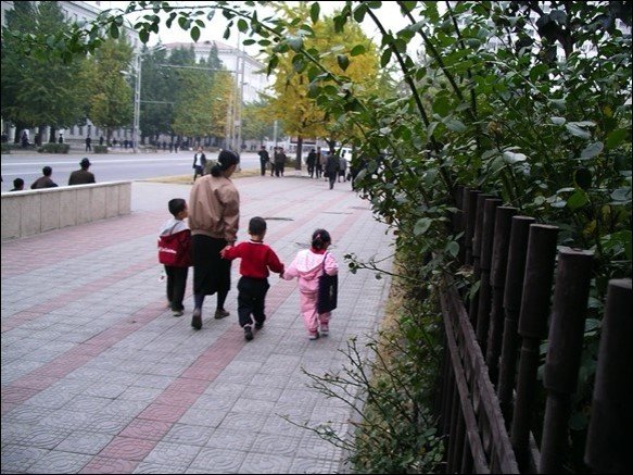 View from the back, woman leading three small kids on a wide nicely paved sidewalk