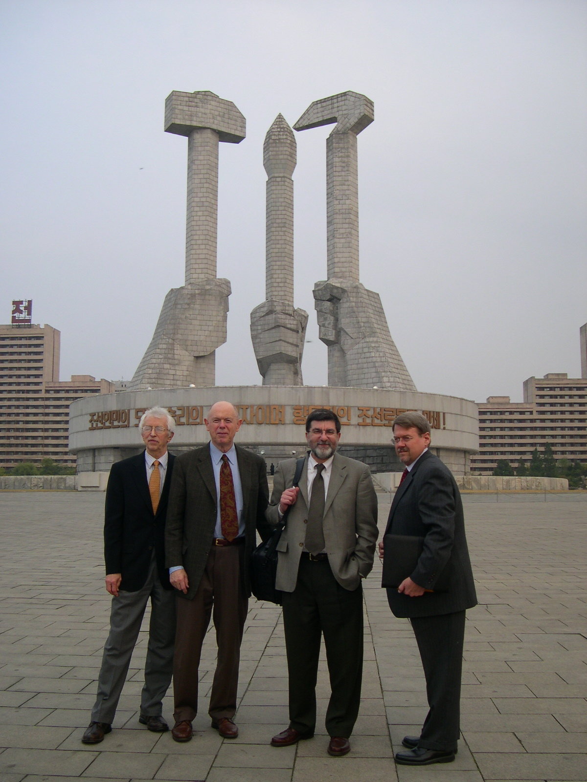 Four men in business suits posing for picture in front of a massive sculptural composition featuring stone fists clutching items symbolizing values of labor and knowledge