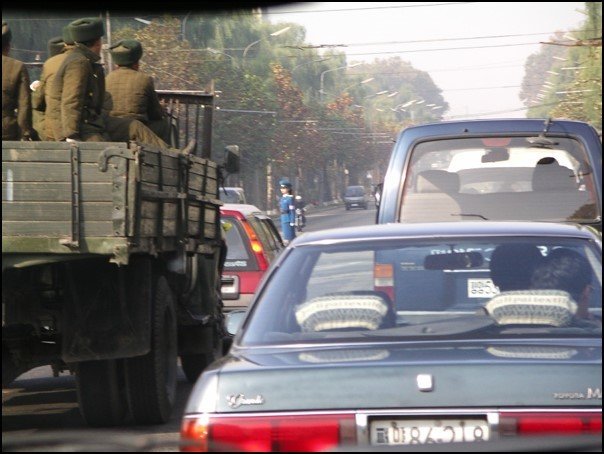 Taken from the front side of a car, backs of cars in front, a truck with some people in military uniform, and a woman traffic controller ahead directing traffic
