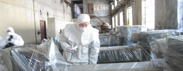 Man in white protective hazmat suit leans over a disabled piece of machinery wrapped in plastic