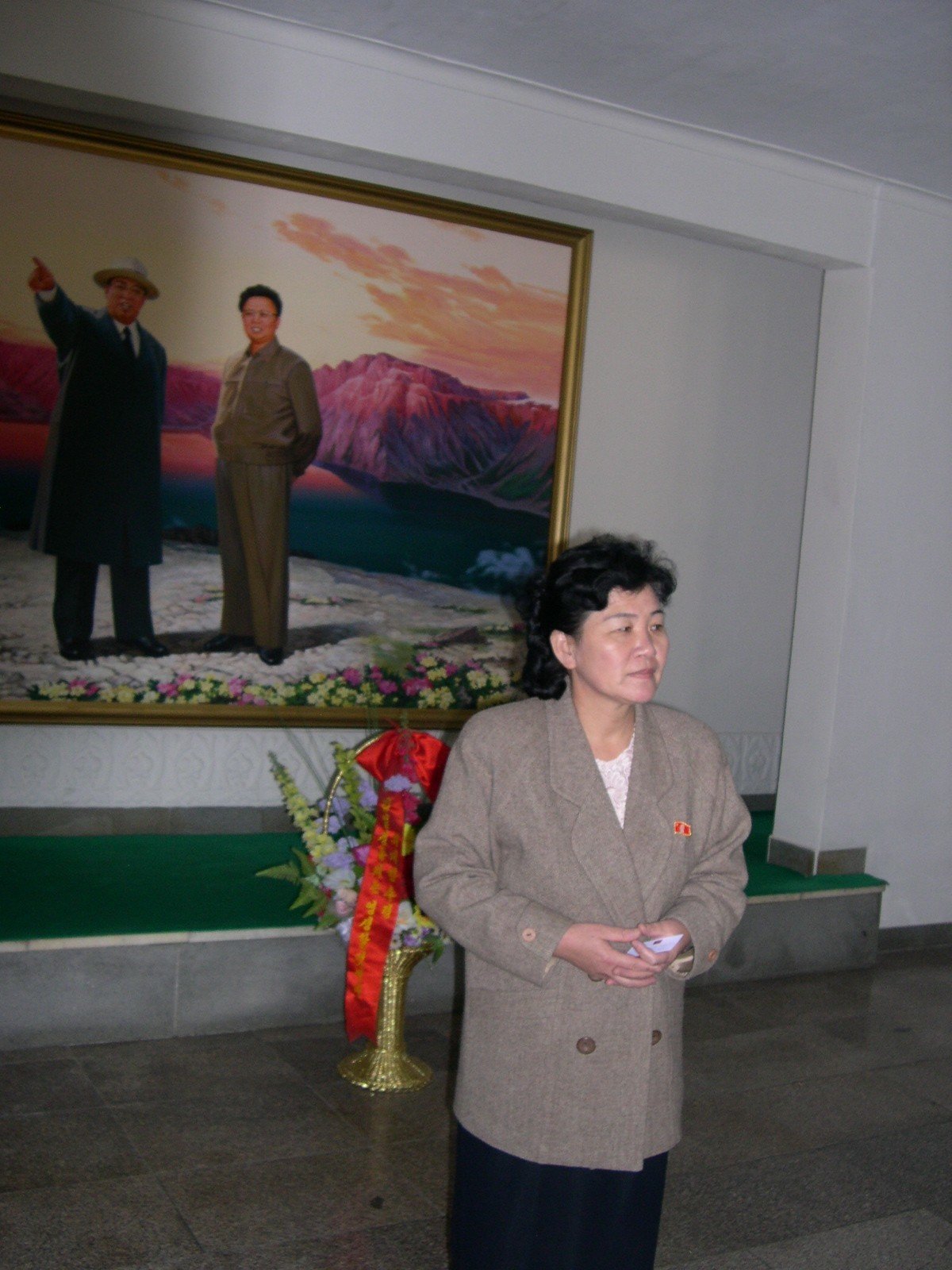 Korean woman in skirt suit is standing in front of a large wall painting of two North Korea leaders