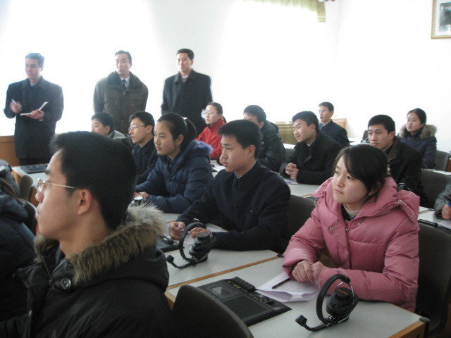 Students in their warm coats seated at desks in a cold classroom and listening attentively to someone outside of the picture. Three men standing at the window looking on.