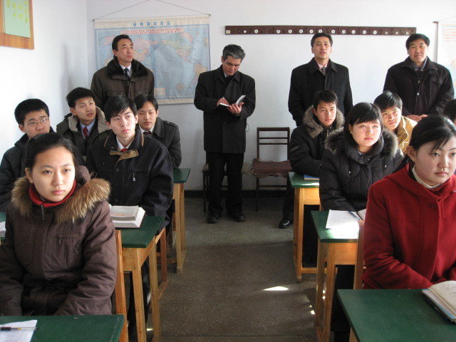 Classroom with warmly dressed students seated in pairs at desks and four men standing in the back of the room
