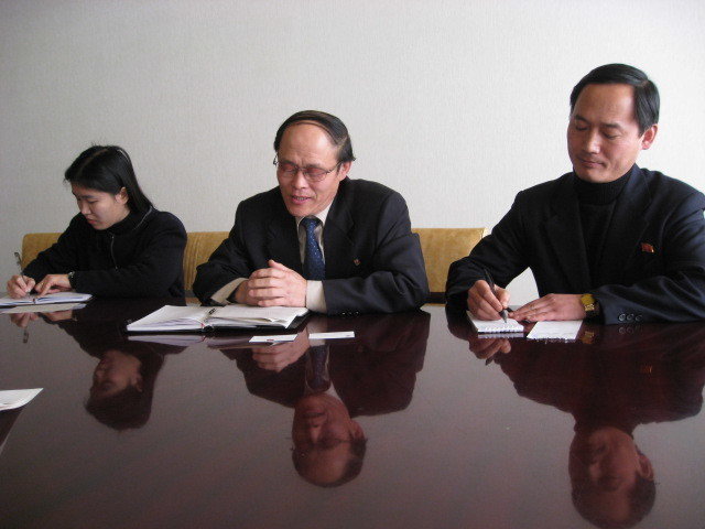 Two men and a woman at the far end seated at a large polished surface desk with their notes at a meeting