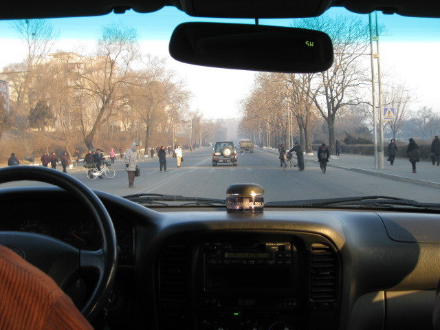 Taken through the car windshield, a city street with a few vehicles and many pedestrians crossing the road on a cold winter morning