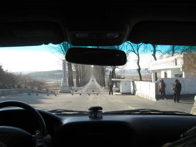 Taken from inside the car, a guard booth and station on the right with a view to a straight free lined valley ahead behind the closed gate