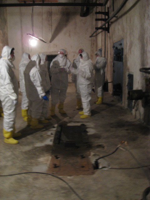 Group of people in anti-c suits talking under a bare electric bulb in a vast industrial space