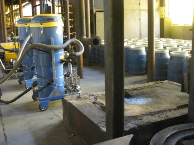 Two tall blue tanks on wheeled platforms with hoses attached and many barrels stored on the floor