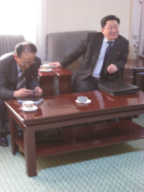 Two Korean men in suits and ties in low armchairs; one looking in his phone, the other leaning forward with a smile