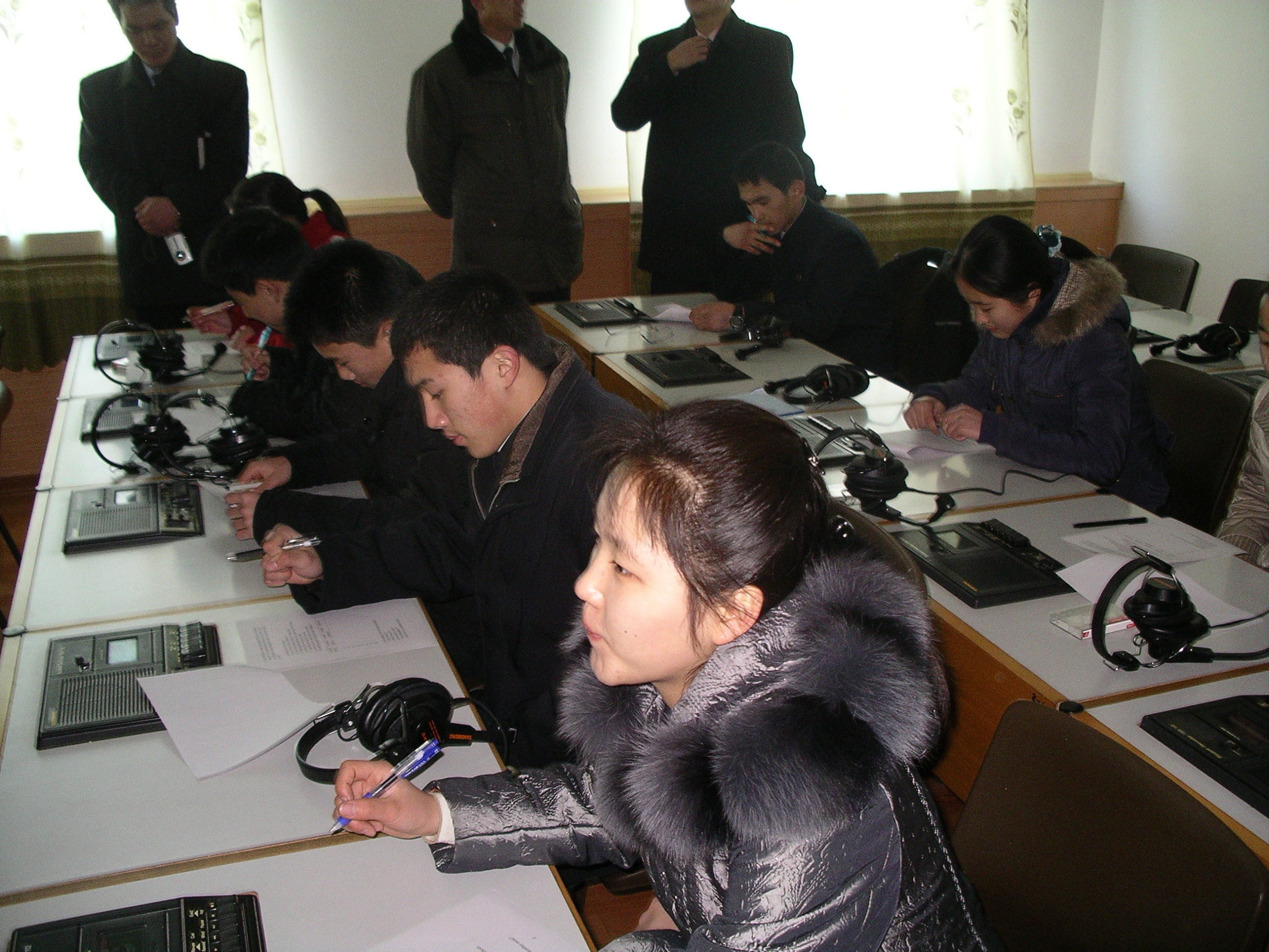 Girl in a winter coat seated at desk with more students seated further in a row with headphones and notebooks in front of them