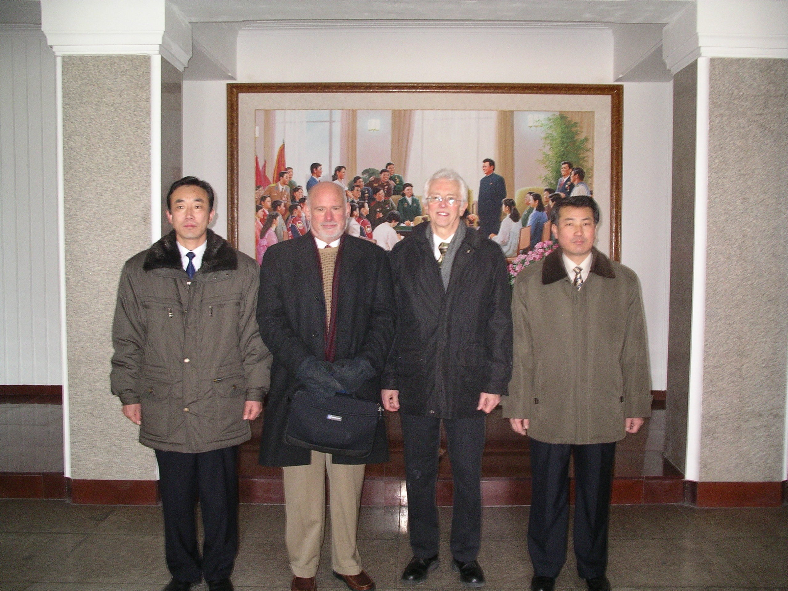 Two Americans flanked by two Koreans all wearing winter coats posing for photo in front of a painting of DPRK leaders