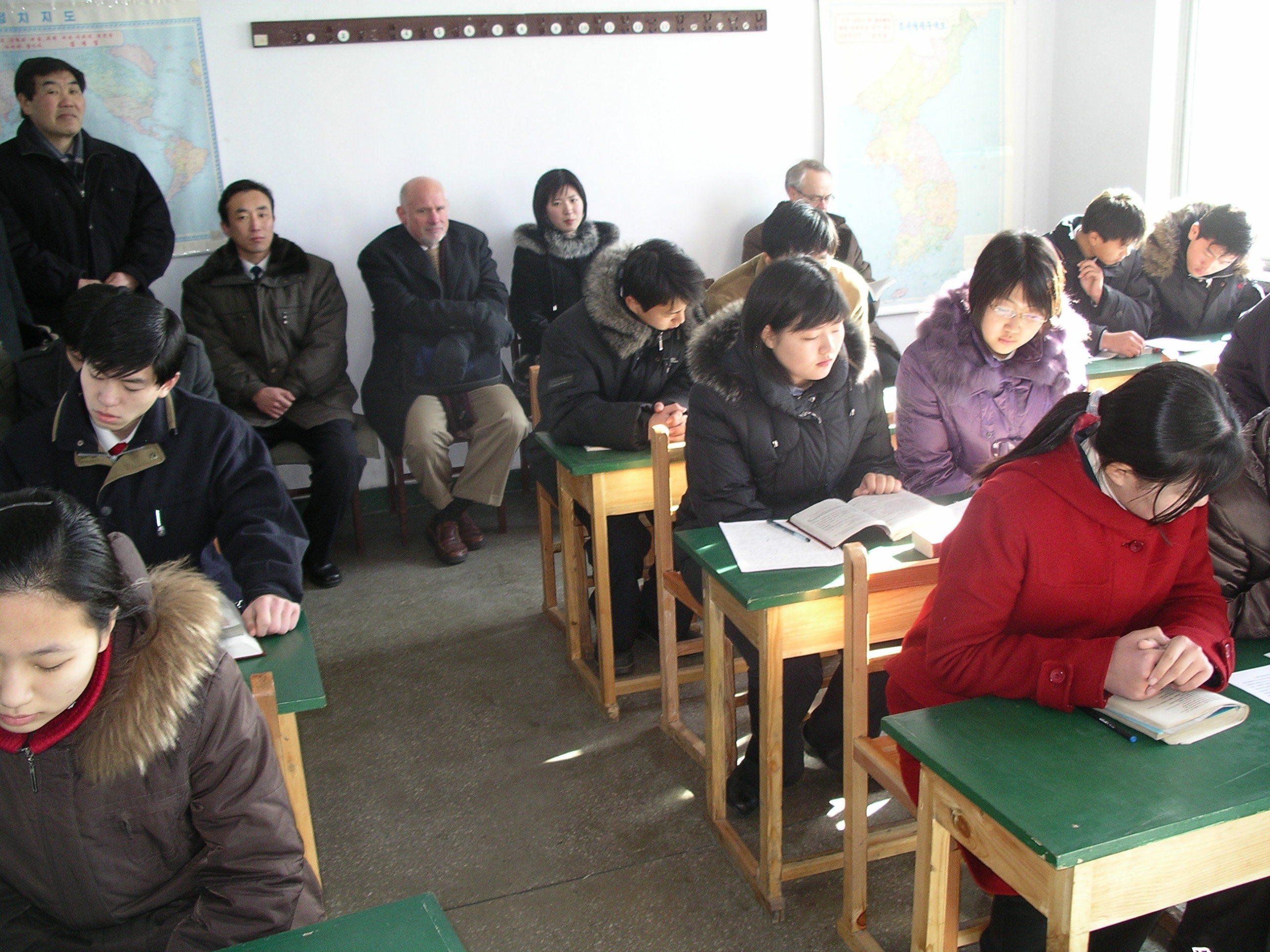Classroom with warmly dressed students seated in pairs at desks. Visitors who look cold seated on chairs in the back of the room
