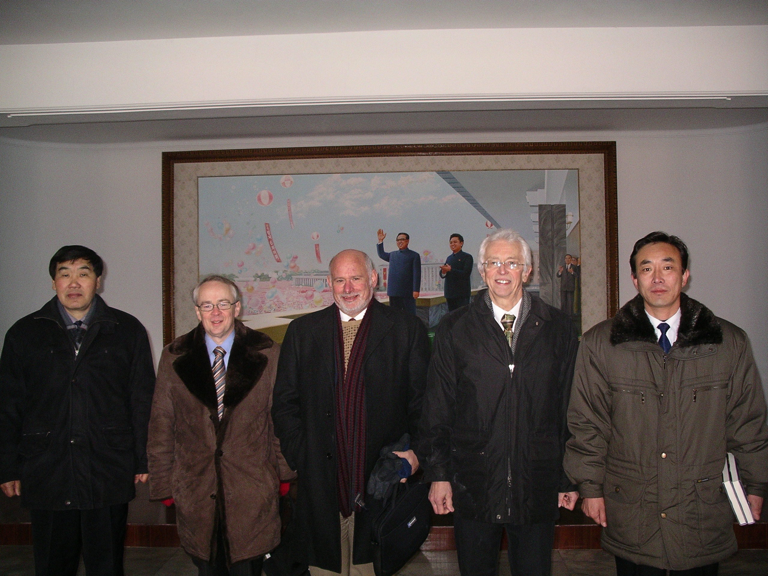 Three Americans flanked by two Koreans all wearing winter coats posing for photo in front of a painting of DPRK leaders
