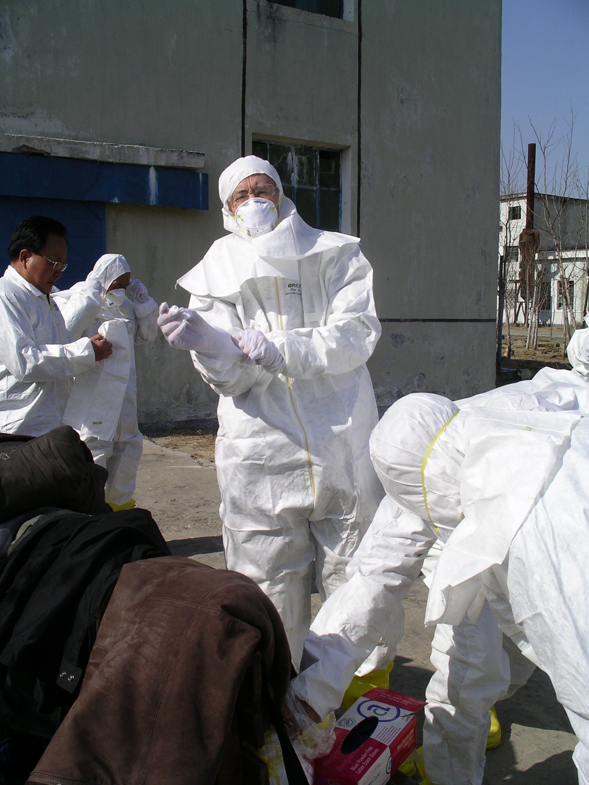 Man donning protective gloves and already in full anti-c suit standing outside with other people adjusting their suits nearby
