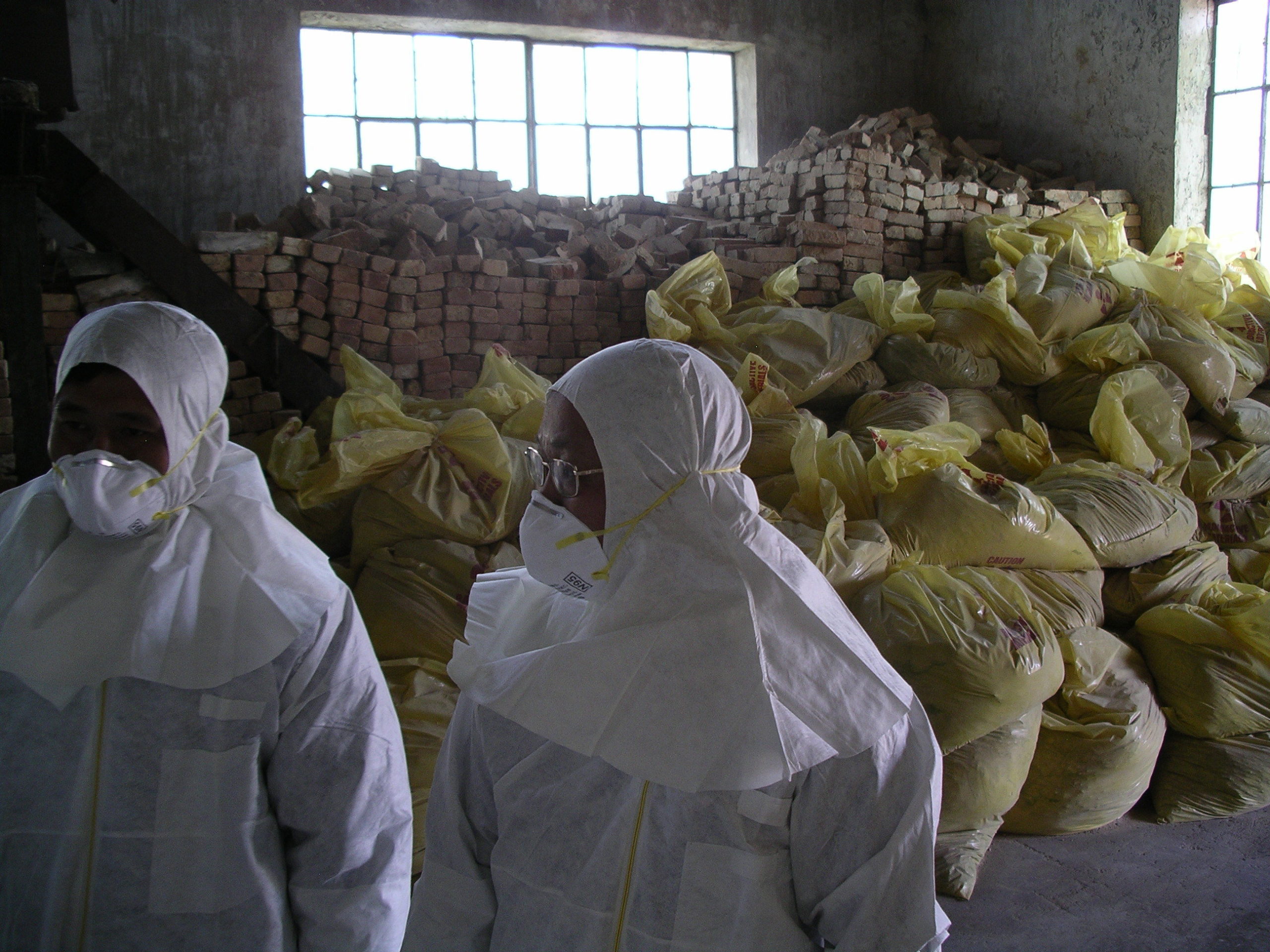 Two men in protective white suits and respirators with heaps of plastic bags and staked bricks in the back