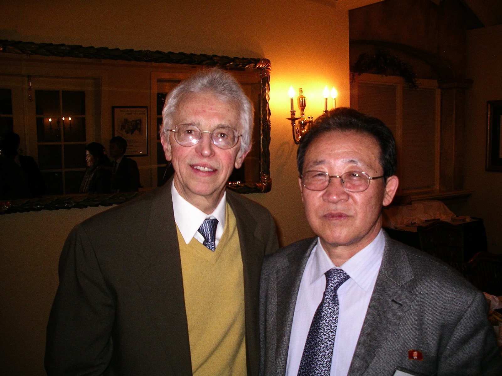 Man in mustard color vest and man in gray business suit both wearing glasses pose for a photo in a restaurant room