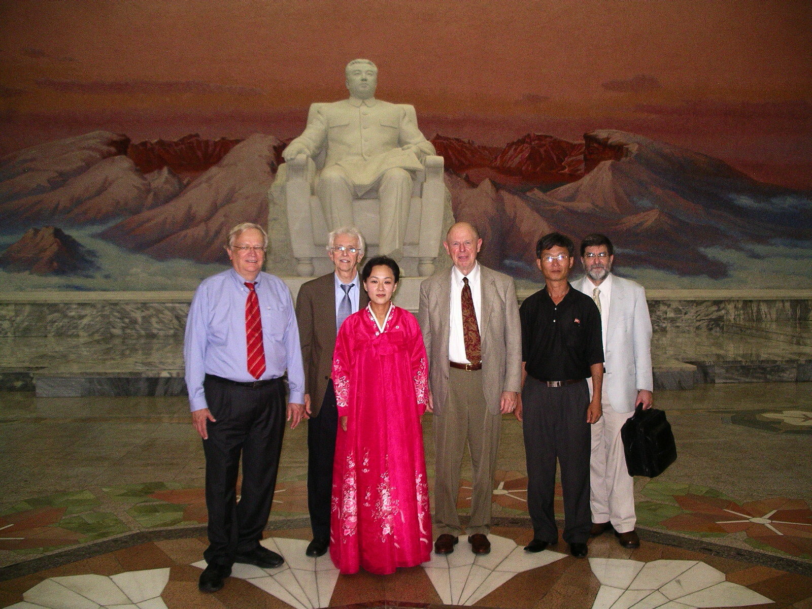 Five men and a woman in pink long Korean traditional gown in center with a seated statue of Kim Il Song behind them