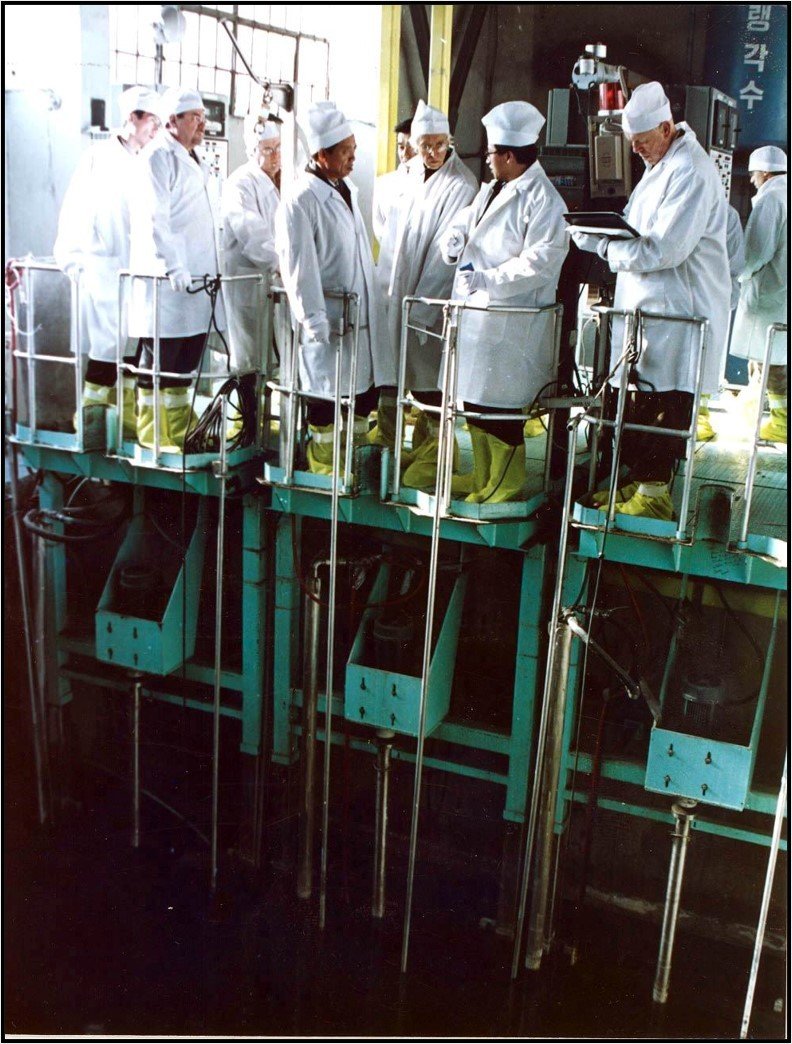 Large group dressed in white robes, caps, and protective footwear, standing on a platform looking down to a spent fuel pool