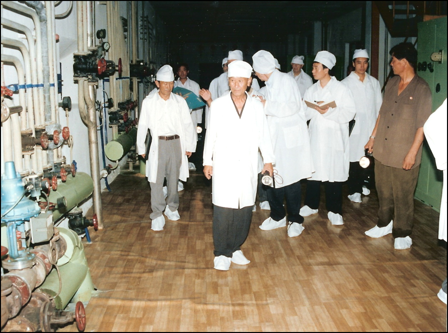 People in white gowns, caps, and booties walking in tight group in a corridor lined with complex piping