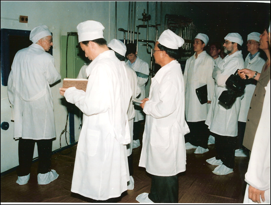 People in white gowns, caps, and booties watching as Hecker peeks into a window in a wall