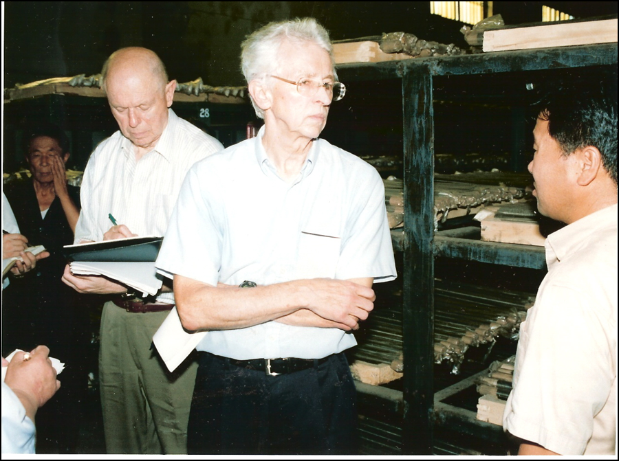 Hecker in white short sleeve shirt standing with his arms folded next to storage racks with uranium rods. Lewis taking notes standing behind.