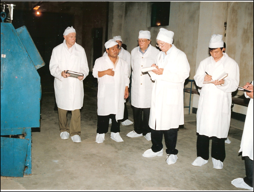 People in white gowns, caps, and booties with notepads listening to a person standing in a large industrial space