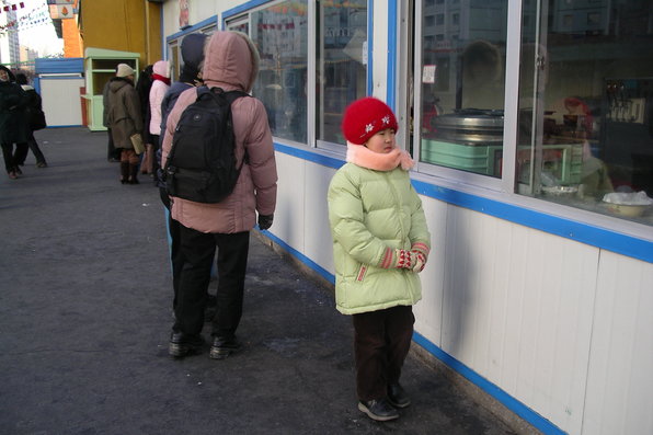 Small kid in bright winter coat and beanie hat looking sideways inside a street food kiosk