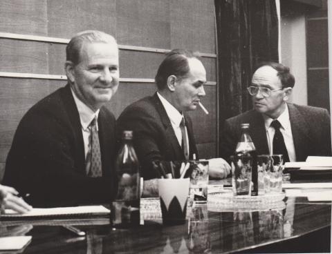 Black and white photo of three men in suits (one smoking) at a panel table
