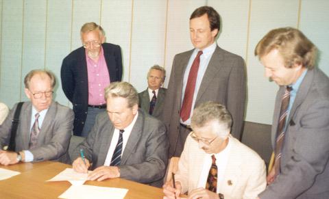 Three people signing documents with men standing behind them