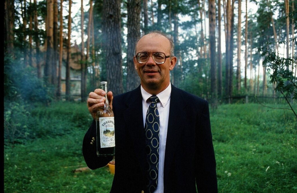 Man in a suit, standing in a wooded area holding a bottle.