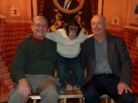 Two men seated with a chimpanzee in between them. The chimp is dressed in a white sweater, jeans, and grey striped socks.