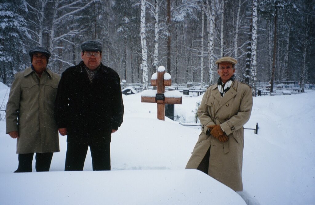 Three men at a snowy grave with crosses behind them.